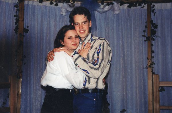 Susan Powell with short hair, wearing white blouse and black jeans embracing Josh Powell wearing a striped Native American-print button-up shirt. Both are standing in front of a backdrop of crepe curtains and ivy.